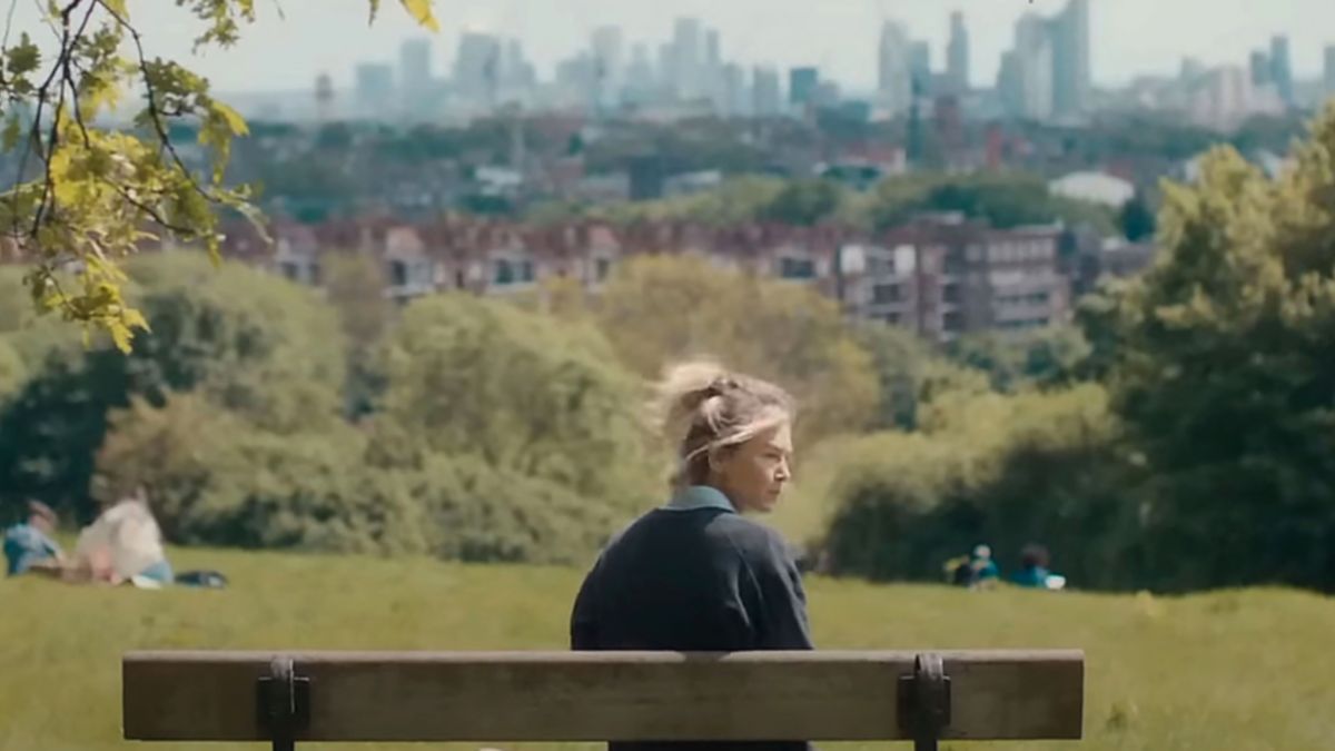 Bridget Jones sitting on a bench in Hampstead Heath looking out across the city of London.