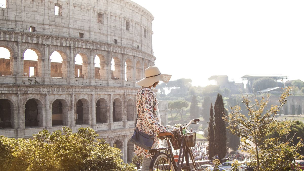 Young adult in fashion dress walking on hill with her bike in front of colosseum Rome.