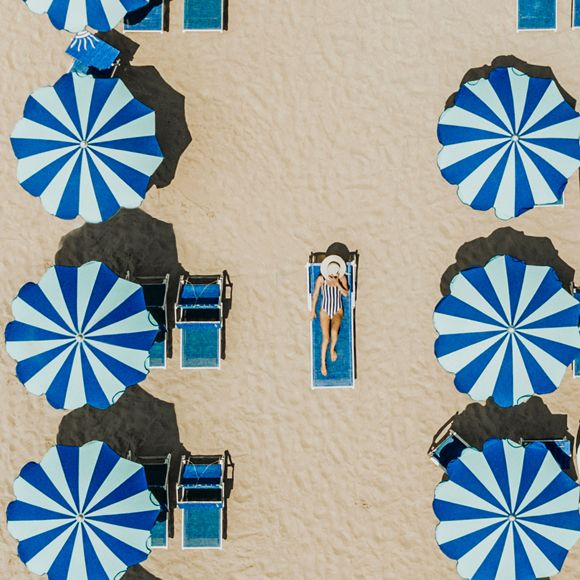 Aerial view over a woman relaxing on the beach surrounded by blue and white striped umbrellas, linking to BA High Life website.