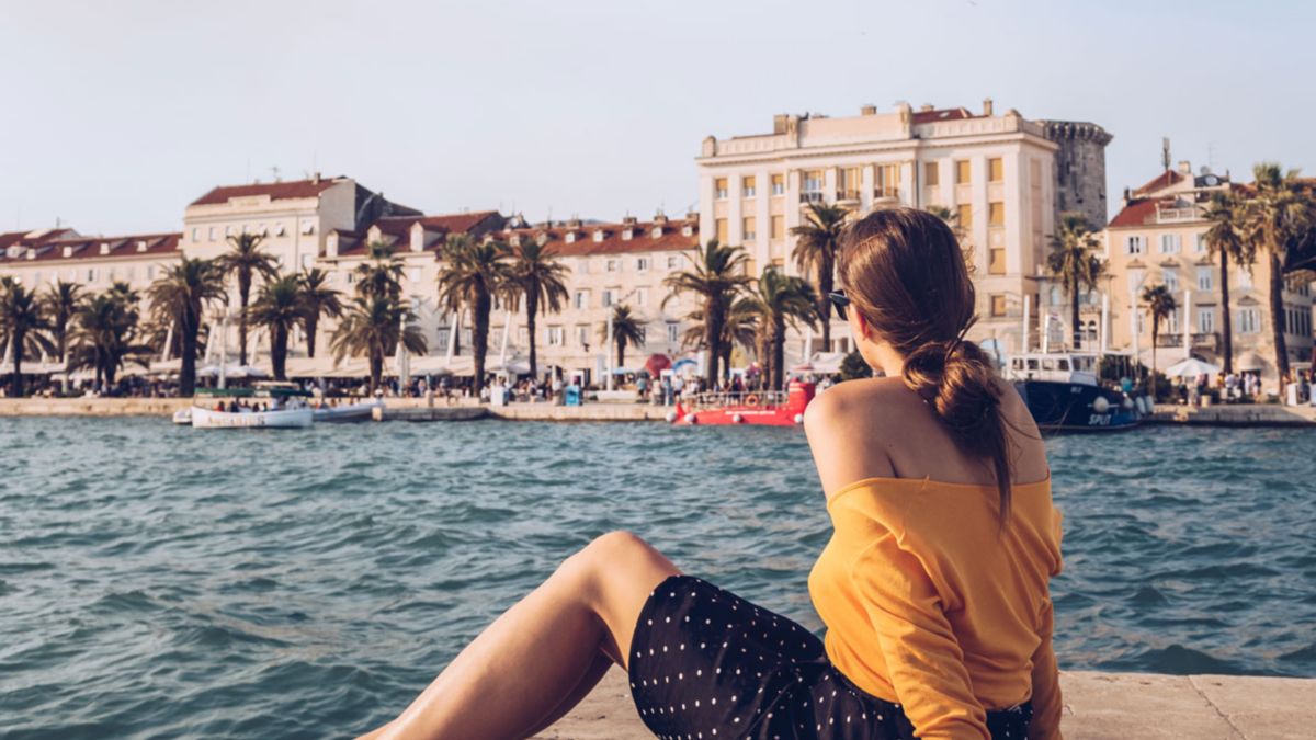 A woman sitting at the waterside looking out across the Split Riva promenade, linking to BA High Life site.​