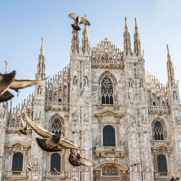 The Gothic architecture of Milan Cathedral with several pigeons flying in the foreground, linking to BA High Life site.