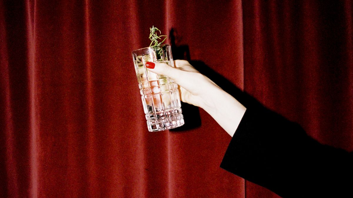 Woman holding cocktail in front of a red velvet curtain, links to BA High Life.com.